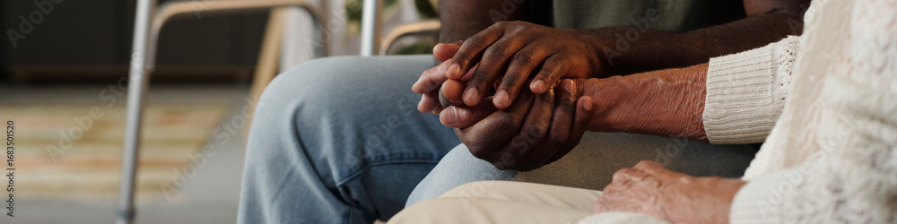 Fototapeta premium Black man holding hands with senior Caucasian woman, offering comfort and support, closeup of intertwined hands showing intergenerational connection and emotional care