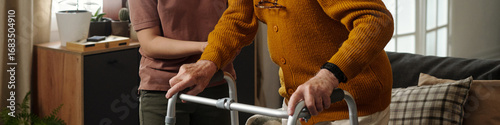 Senior Caucasian man using walker for support while young adult woman assisting him indoors, both standing close together, partial view focusing on upper bodies and hands