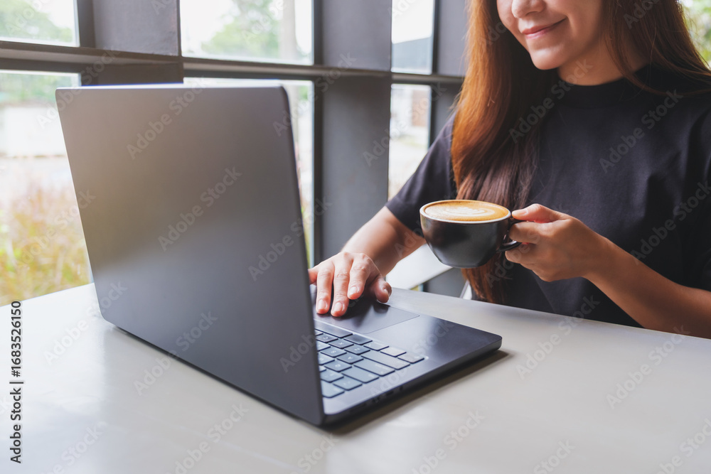 Fototapeta premium Closeup image of a woman working and typing on laptop computer while drinking coffee in cafe