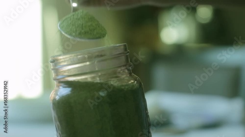 Close-up view of green herbal salt being poured from a spoon into a glass jar. Concept of healthy seasoning, organic food, and natural culinary ingredients.
