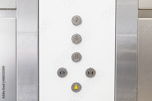 Elevator control panel with metallic buttons and yellow alarm bell. Closeup of floor selection and door operation keys in modern lift interior. Minimal design of stainless steel elevator wall.