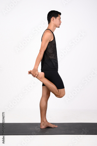 A male runner performs a standing quadriceps stretch on a yoga mat, balancing on one leg while pulling his heel toward his glute.