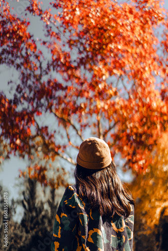 A woman with long hair wearing a brown hat and a patterned coat. She is standing under the vibrant red autumn leaves of the maple in Thailand. Idea for traveling wallpaper.