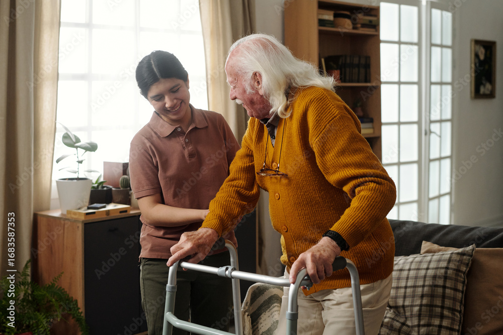 Fototapeta premium Young adult Latin woman assisting senior Caucasian man using walker in living room, both smiling and interacting, caregiver supporting elderly patient during rehabilitation activity