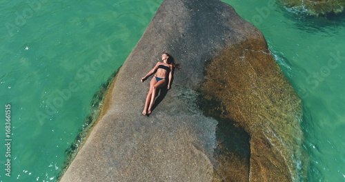 A person enjoys sunbathing on a large rock surrounded by vibrant turquoise water. The scene showcases a serene atmosphere perfect for relaxation on a warm day, highlighting natural beauty.