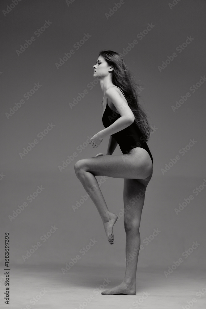 Fototapeta premium Black and white photo of a young woman in a leotard posing with raised leg, showing elegance, grace, and balance in studio lighting.