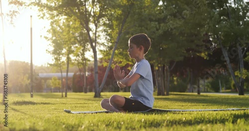 Child boy practices meditation in lotus pose on yoga mat in summer park.Calm kid with closed eyes and folded hands enjoys mindfulness, breathing and harmony with nature.Concept of wellness and balance