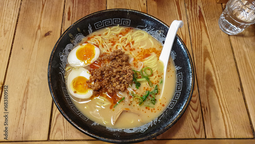 Photography A delicious bowl of spicy Tantanmen ramen with a creamy broth, savory ground pork, scallions, and a perfectly soft-boiled ajitama egg, served on a rustic wooden table in a Tokyo noodle shop