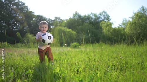Young boy is holding soccer ball. Male child holds a soccer ball outdoors. A casual field of greenery and grassy nature in summer. A young boy is grasping a soccer lifestyle ball.