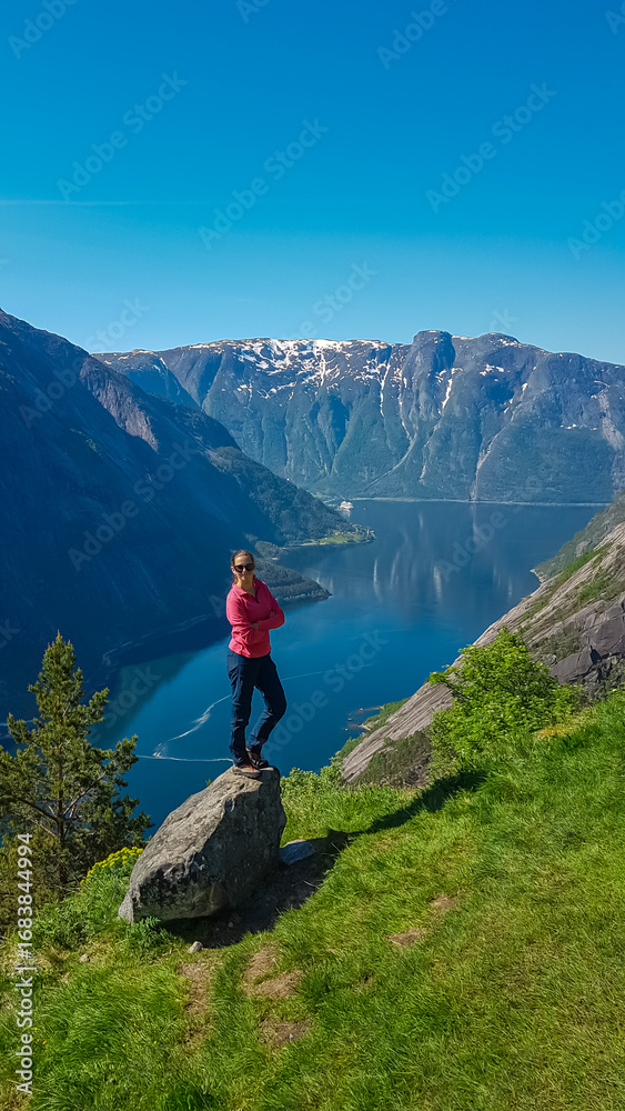 Naklejka premium A triumphant female hiker stands confidently on a rock at the Kjeåsen viewpoint, overlooking a Norwegian fjord. Her pose against the vast mountain landscape celebrates adventure and outdoor travel.