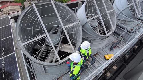 Two workers perform maintenance on large cooling fans situated on the rooftop of a commercial building in a bustling urban environment. They are dressed in safety gear and inspecting equipment.