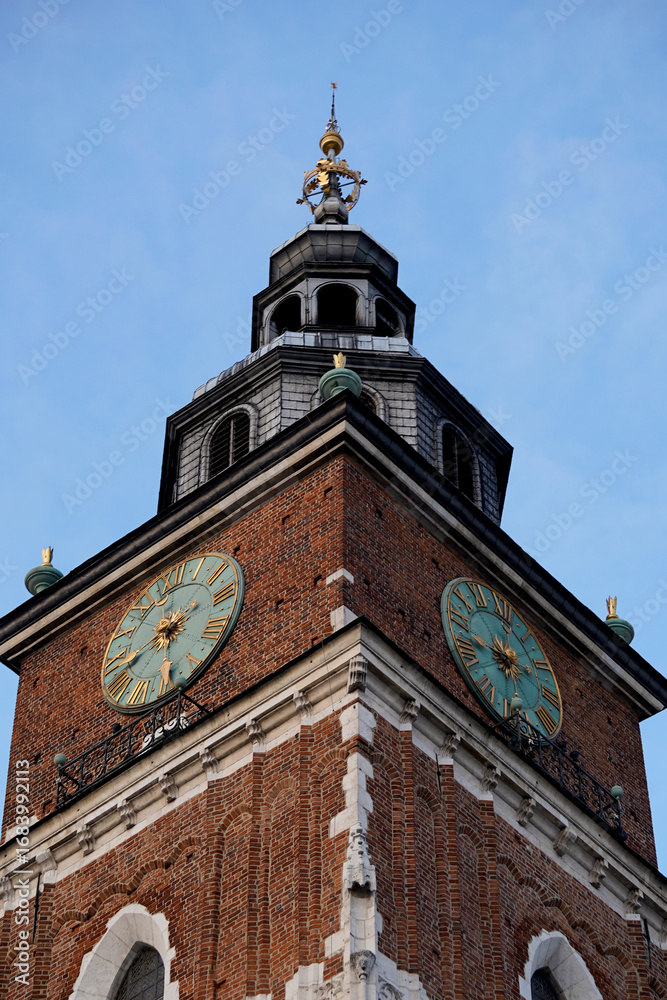 Obraz premium Historic brick clock tower with green dials and ornate spire rises against clear blue sky, symbolizing tradition, time, and architecture