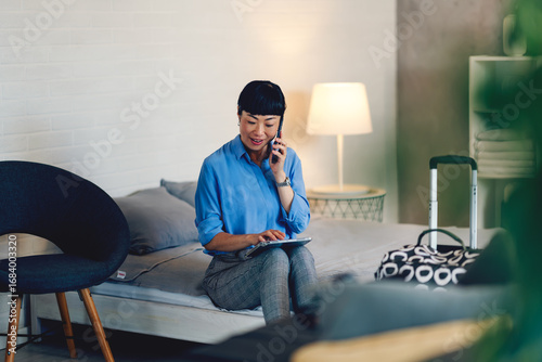 Photos Smiling middle-aged Asian woman sitting on hotel bed, talking on smartphone and