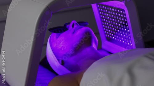 Woman with hydrogel mask on face in protective glasses getting violet LED light facial therapy treatment in beauty salon, laying on couch, relaxing. Beauty, rejuvenation, anti-aging, wellness concept
