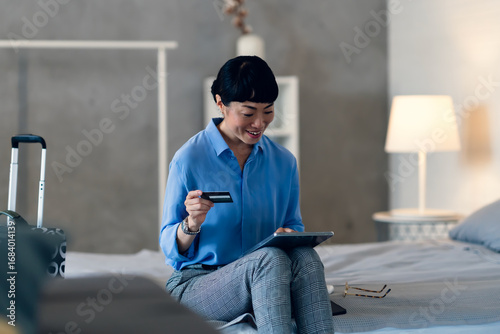 Tableau sur toile Smiling middle-aged Asian woman sitting on hotel bed with tablet, holding a cred