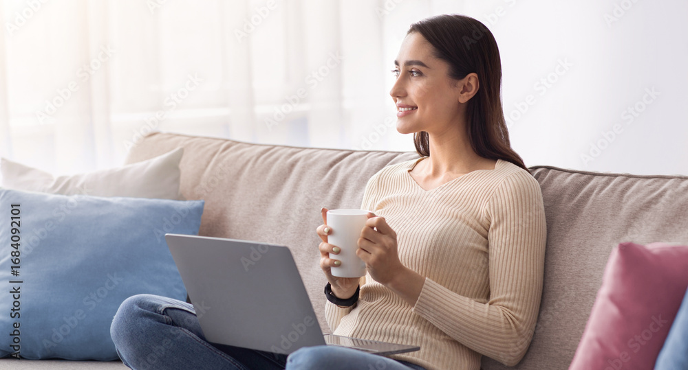 Naklejka premium Enjoying free time and social networks at home. Beautiful young muslim woman work on laptop and drinking coffee. Cheerful pensive arab lady on sofa looking towards empty space in living room interior