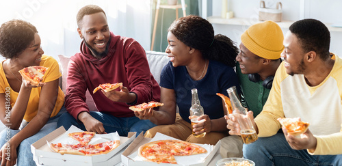 Foto Joyfull african american millennial friends eating pizza in living room, drinkin