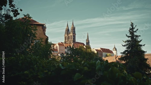 View of Albrechtsburg Castle and Meissen Cathedral rising above the old town rooftops, framed by greenery under a clear summer sky in Saxony, Germany.