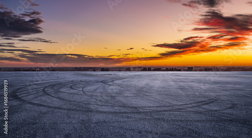 Empty asphalt race track ground with car tire skid marks and city skyline at dramatic sunset