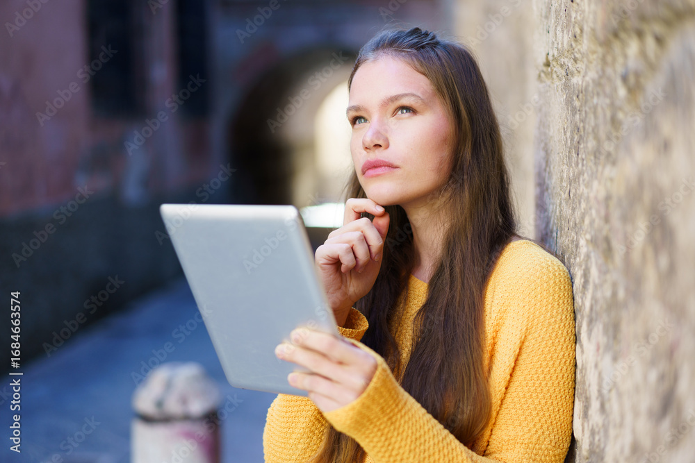 Obraz premium A Thoughtful Young Woman Engaged with Her Tablet While Sitting in an Urban Environment