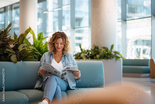 Free Download Smiling woman in denim outfit relaxing on a modern sofa while reading a magazine in a bright, plant-filled indoor space - GFXLoad