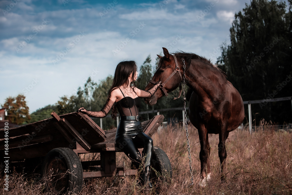 Obraz premium Sexy beautiful young woman in leather clothes petting a horse while sitting on wooden vintage cart in the field. Freedom and imprisonment. Friendship and loyalty. Peace of mind. Dramatic and nostalgic