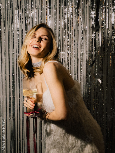 Young blonde woman wearing white dress and necklace is laughing and holding glass of white wine in front of silver curtain during party. Woman in white dress, necklace, laughing, holding wine glass