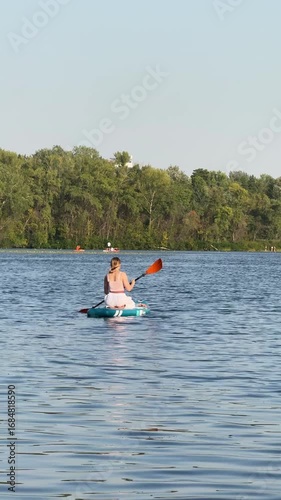 Woman sits on a SUP board paddling along a river with forest in the background at sunset, viewed from behind, enjoying outdoor adventure and calm nature