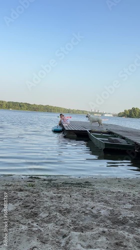 Woman stands with her dog on a wooden pier, launching a SUP into the river at sunset, enjoying a peaceful outdoor moment in nature