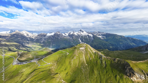 Aerial view of the serpentine roads winding up to the observation point on the lush green mountain, with snow-capped peaks in the distance, Bruck an der GroÃŸglocknerstraÃŸe, Salzburg, Austria.