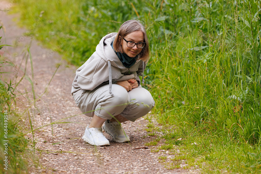 Fototapeta premium Person crouching on a nature path observing plants in a lush green environment during daylight hours