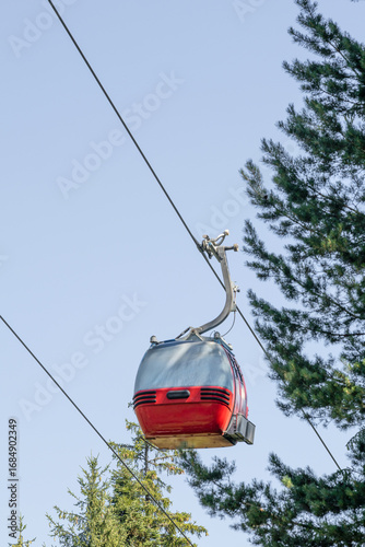 A vibrant red and silver gondola lift cabin is suspended against a clear blue sky, with evergreen trees visible. This iconic transport system is a common sight in the Bansko ski resort.