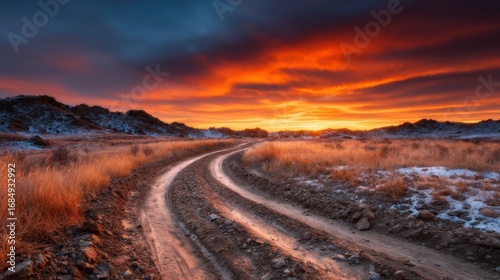 A winding dirt path leads through a striking landscape as a vibrant sunset paints the sky with vivid orange and purple hues, creating a tranquil and scenic atmosphere.