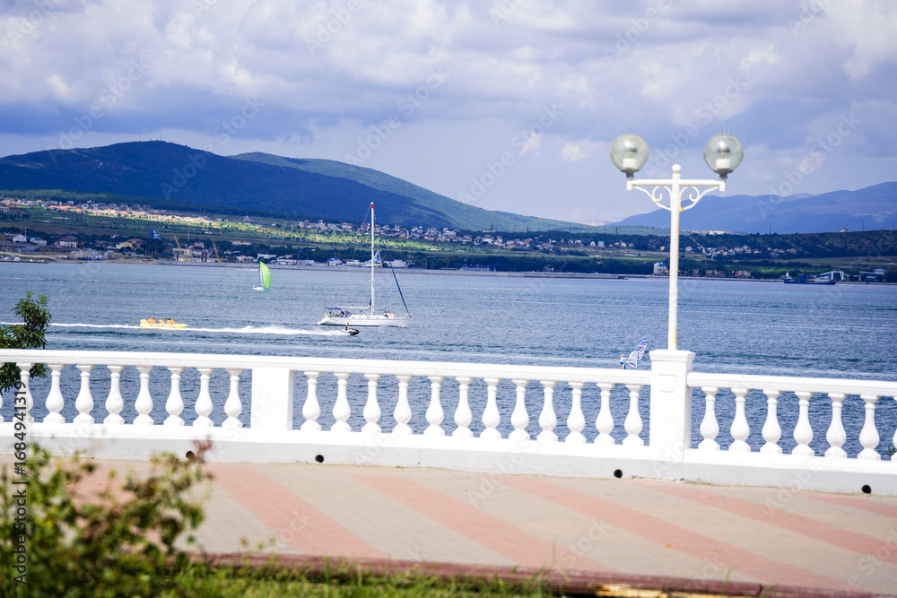 Obraz premium The embankment of the Gelendzhik seaside resort: white balustrade, sea, mountain range in the background. Summer, white clouds