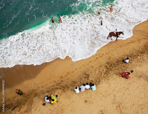 Aerial view of golden sands meet turquoise waves, where people gather and a horseman rides along the shore, Mahabalipuram, Tamil Nadu, India.