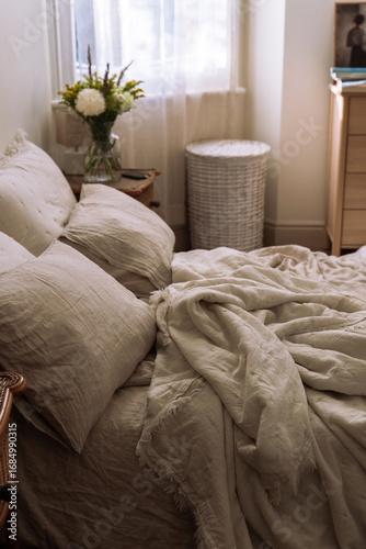 Cozy bedroom scene with oatmeal linen bedding, pillows and a glass vase of fresh flowers on a wicker nightstand, soft natural light.