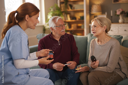 A home health aide is explaining medication dosages to a senior couple at their residence. The nurse is reviewing a pill bottle and blister pack with them as they sit on the sofa in their living room.