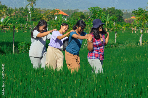 With arms on shoulders, four cheerful girls sing and laugh together while walking along the narrow rice field path surrounded by greenery and sunshine.