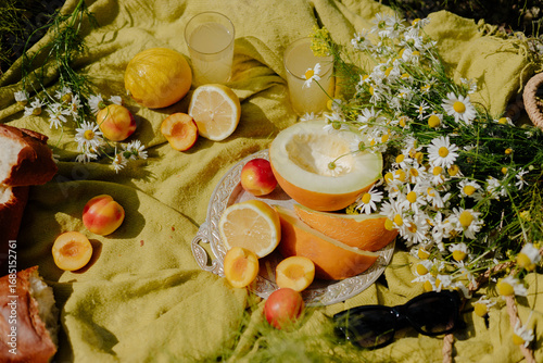 A vibrant picnic setting features fresh fruits like melons, lemons, and apricots, along with wildflowers, showcasing a joyful summer gathering on a sunny day