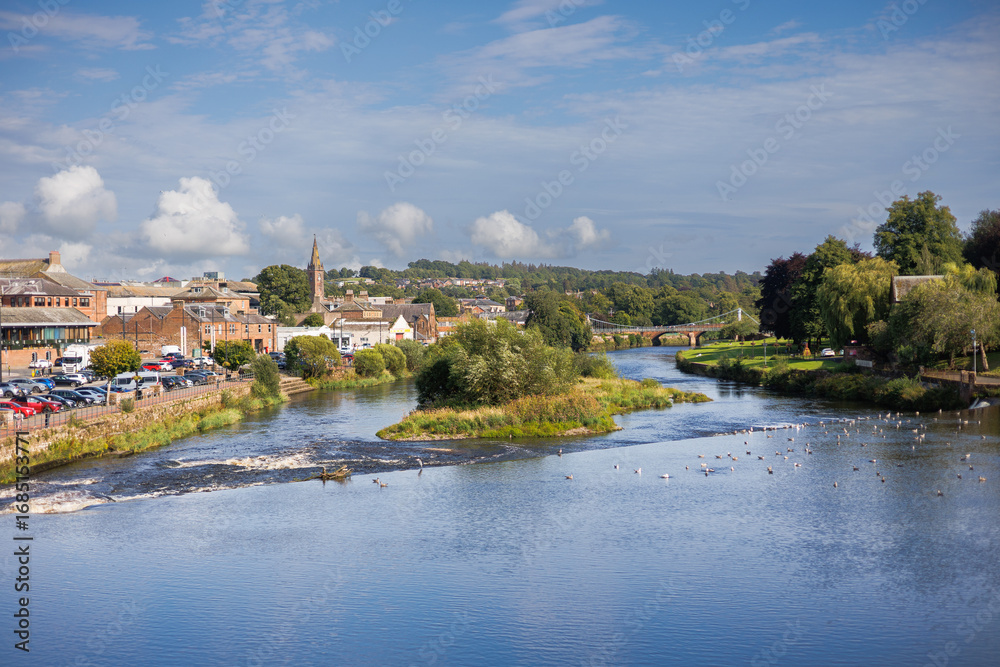 Obraz premium Scenic view of a river flowing through Dumfries, Scotland, taken from a bridge, with reflections and surrounding urban and natural landscape