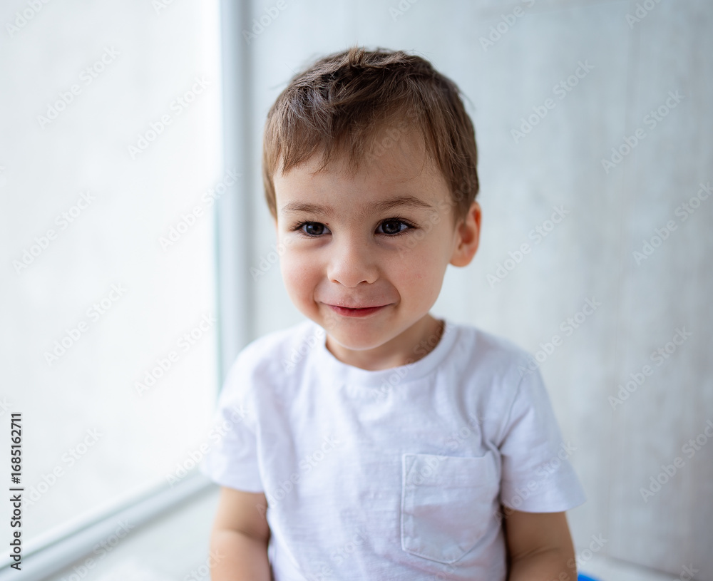 Naklejka premium Smiling boy in casual clothing indoors. A young boy with short brown hair smiles happily in a bright indoor setting, wearing a simple white shirt.
