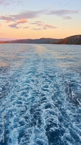 Vertical video: a serene view from the stern of a ship sailing away from the coast at sunset. The vessel sways slightly, creating a meditative mood. Ideal for social media.