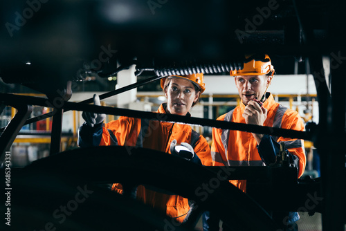 Bild auf Leinwand Two engineers wearing safety gear and helmets inspect machinery under train in railroad maintenance facility, showcasing teamwork, mechanical engineering and railway system inspection