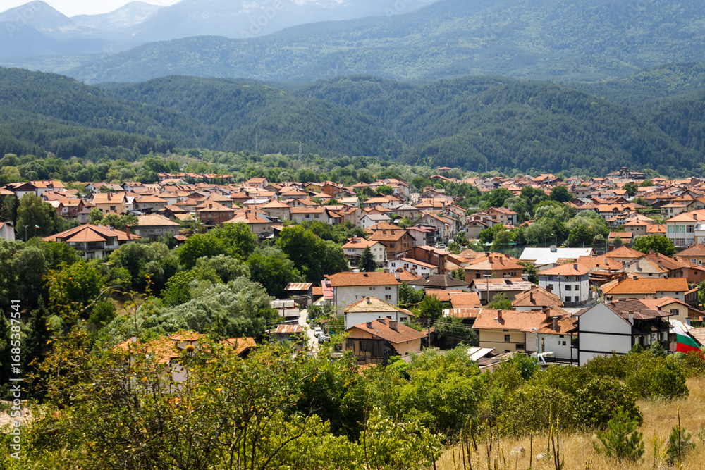 Fototapeta premium A picturesque panoramic view of the Bulgarian town of Dobrinishte, nestled in a valley with its terracotta-roofed houses peeking through lush green trees. Majestic mountains frame the background. 