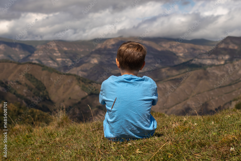 Naklejka premium Rear view of a boy sitting on a rock with mountains in the background