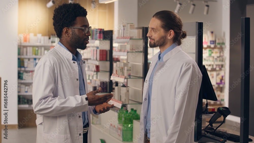 Obraz premium African Caucasian men doctors conversation in pharmacy medicine smiling guys physician pharmacist health care male coworkers in medical coat talk drugstore professional assistance looking at camera