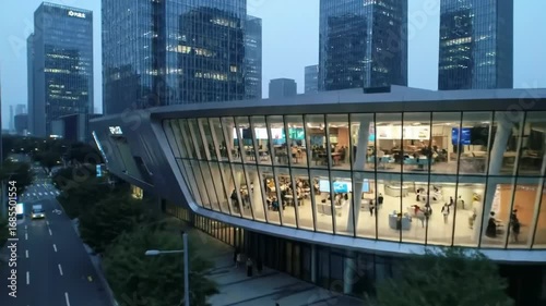 An urban landscape shows a pathway leading towards a modern building with lights glowing in the evening atmosphere.