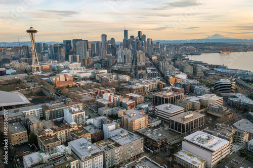 Aerial panoramic view of Seattle waterfront with city skyline, Mount Rainier in the background, Elliott Bay, piers, ferries, modern skyscrapers, trains, and dramatic sunset sky over Puget Sound