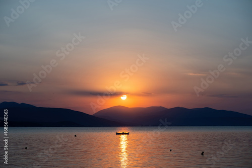 Tranquil fishing boat at sunset over Tolo Bay Greece. Glowing orange sky and distant mountain silhouettes reflecting on calm sea water