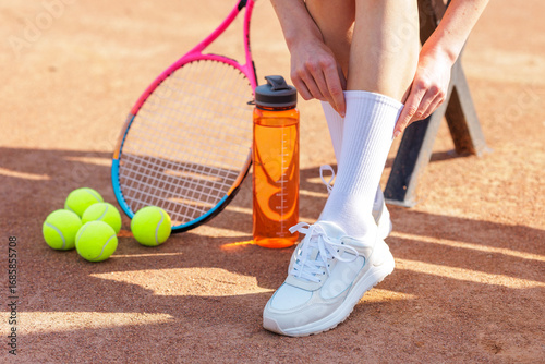 Female tennis player adjusting sock on clay court with racquet balls and water bottle